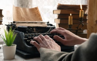 male hands on antique black typewriter with stack of old books in background and a lit candle, mystery pacing