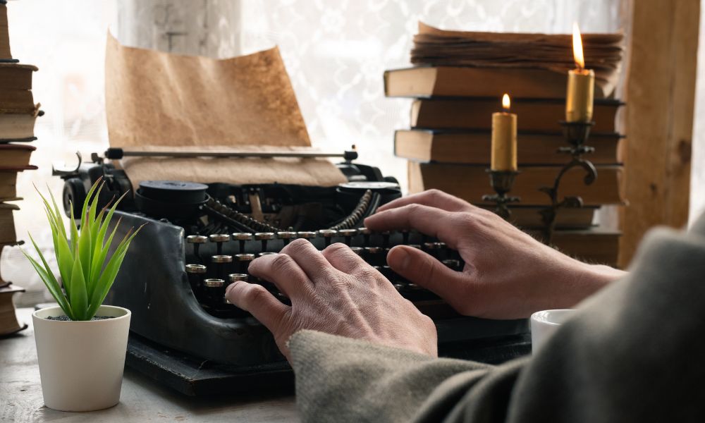 male hands on antique black typewriter with stack of old books in background and a lit candle, mystery pacing