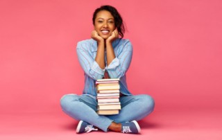woman sitting in front of pink background with a stack of books in her lap; how to create a bookstagram