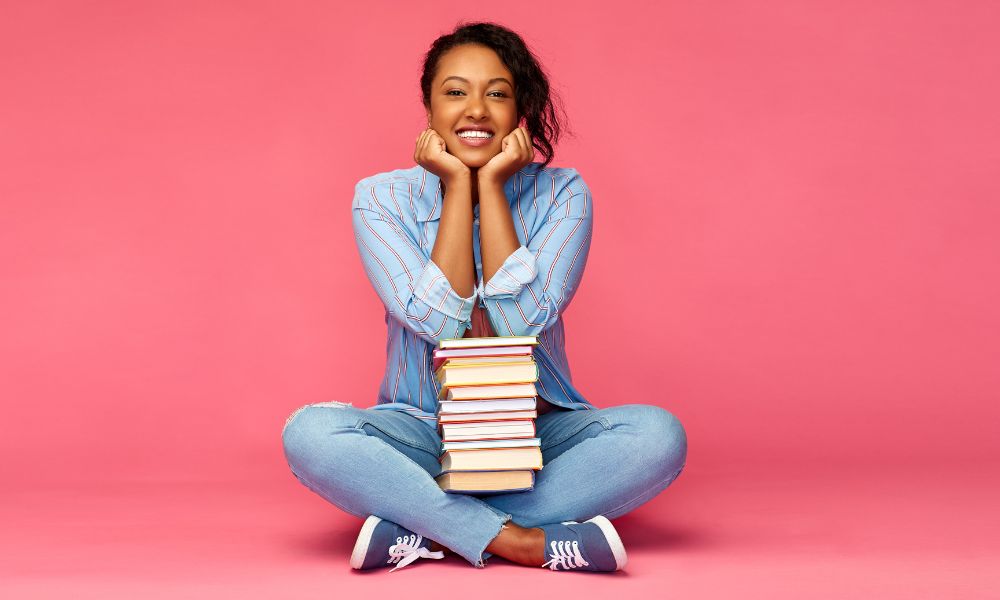 woman sitting in front of pink background with a stack of books in her lap; how to create a bookstagram