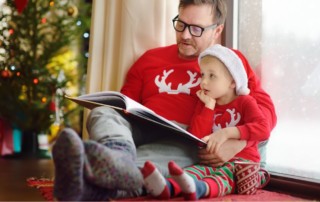 father reading a children's book to a little boy, both are wearing festive holiday pajamas and sitting in front of a Christmas tree; how to write holiday-themed children's books