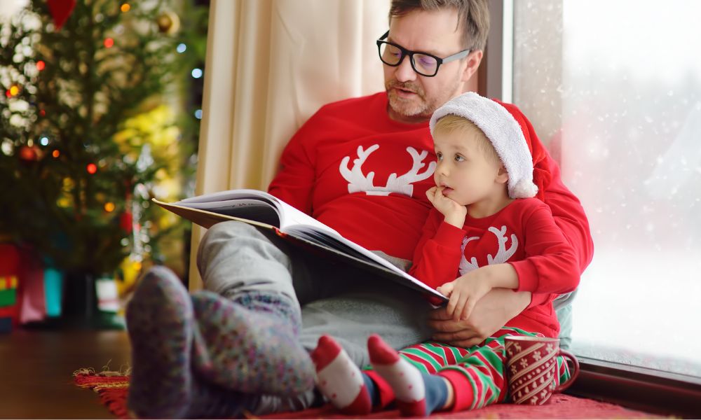 father reading a children's book to a little boy, both are wearing festive holiday pajamas and sitting in front of a Christmas tree; how to write holiday-themed children's books