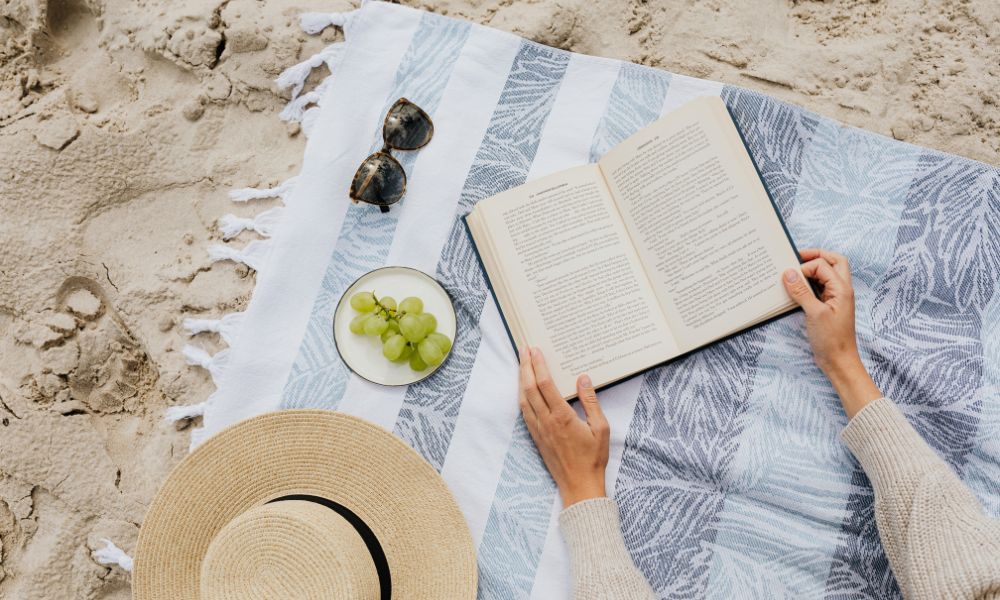 striped blanket on the sand, female hands holding a book open and sunglasses, green grapes on a plate, and a sun hat laying on the blanket; advanced reader copies