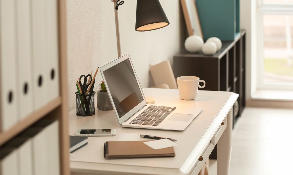 Apple laptop sitting open on a white desk, there is a desk lamp turned on and office accessories on the desk; Mill City Press Editorial Critique