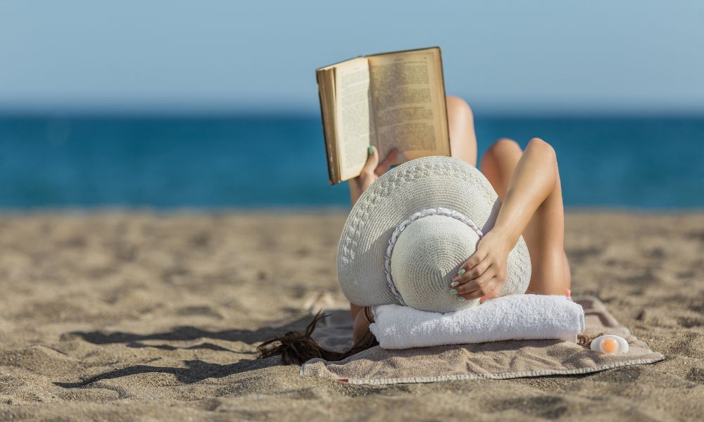 woman laying on a beach towel reading a book at the beach; beach reads